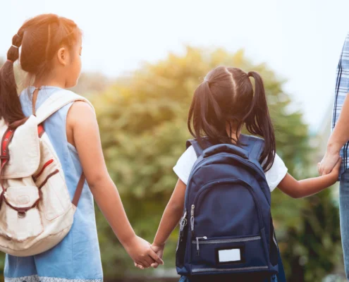 Two school aged girls holding hands waiting for the school bus with their backpacks on and father.