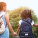 Two school aged girls holding hands waiting for the school bus with their backpacks on and father.